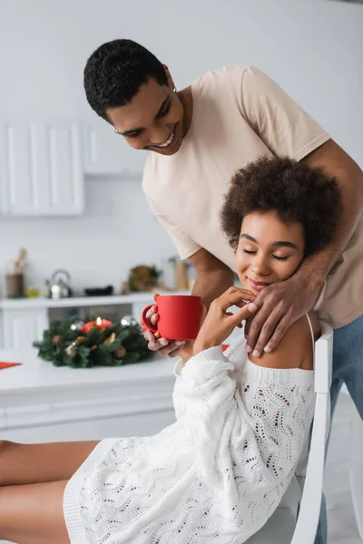 Smiling african american man with red cup touching seductive girlfriend smiling with closed eyes in kitchen — Stock Photo