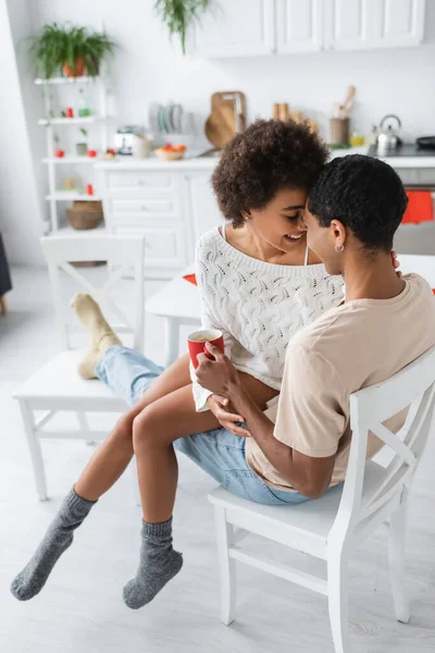 Sexy african american woman in warm socks and white openwork sweater sitting on one chair with boyfriend in kitchen — Stock Photo