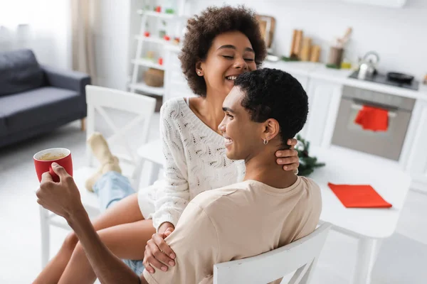 Happy african american man holding cup while sexy girlfriend sitting on his laps in kitchen — Stock Photo