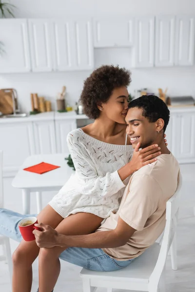 Sensual african american woman hugging boyfriend sitting in kitchen with red cup — Stock Photo