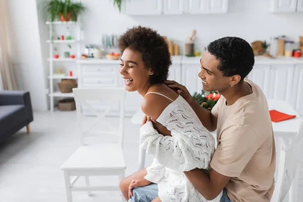 Giovane africano americano uomo abbracciare fidanzata in bianco traforato maglione ridere in cucina — Foto stock