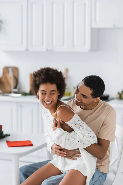 Africano americano homem sentado na cozinha e abraçando rindo namorada em suéter branco openwork — Fotografia de Stock