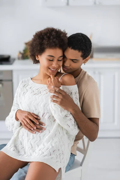 Young african american man sitting on kitchen chair with sexy girlfriend in openwork sweater — Stock Photo