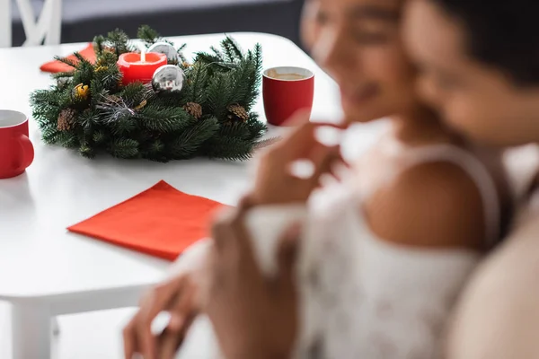 Foyer sélectif de couronne de Noël avec bougie près des tasses rouges et le couple afro-américain sur le premier plan flou — Photo de stock