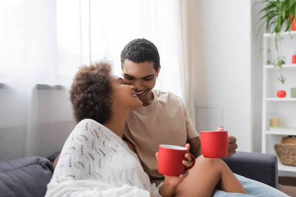Sexy afrikanisch-amerikanische Frau im weißen durchbrochenen Pullover und glücklicher junger Mann sitzt auf der Couch mit roten Tassen — Stockfoto