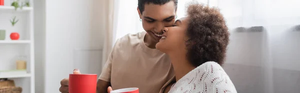Joven y alegre pareja afroamericana sosteniendo tazas rojas y besándose en casa, pancarta - foto de stock
