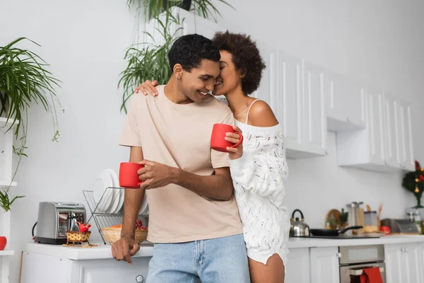 Щаслива афроамериканська жінка в білому светрі тримає червону чашку і цілує чоловіка на кухні — стокове фото