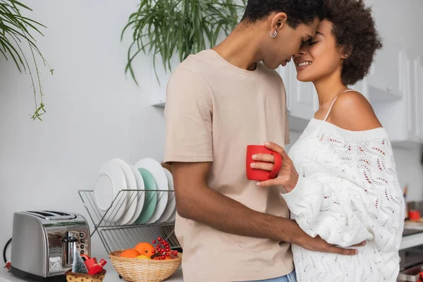 Feliz casal afro-americano abraçando na cozinha perto de torradeira e cesta de vime com laranjas frescas — Fotografia de Stock