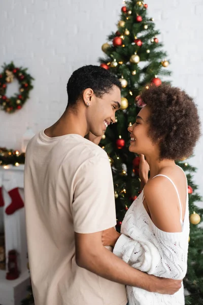 Joven africano americano pareja sonriendo el uno al otro cerca decorado árbol de Navidad - foto de stock