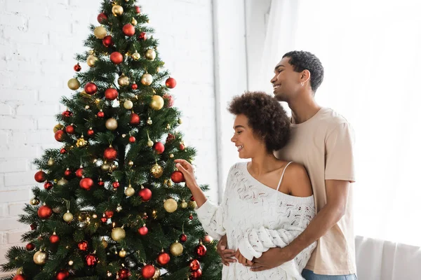African american man hugging girlfriend in white openwork sweater decorating pine tree with christmas baubles — Stock Photo