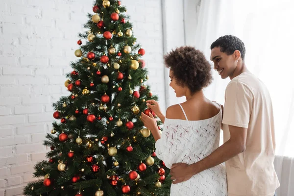 Усміхнений афроамериканський чоловік обіймає сексуальну жінку, прикрашаючи ялину різдвяними вадами — стокове фото