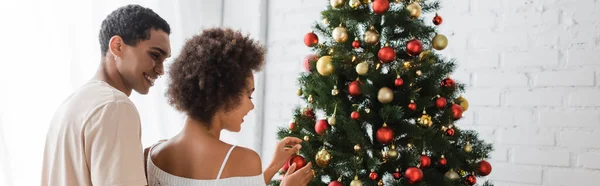 Cheerful african american man looking at curly girlfriend decorating christmas tree at home, banner — Stock Photo