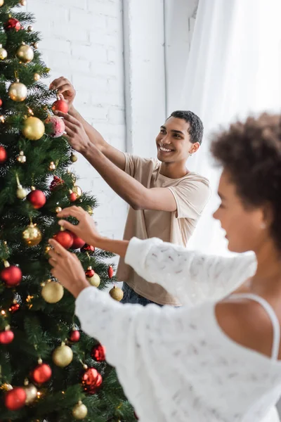 Счастливый африканский американец украшает рождественскую елку рядом с подругой на размытом переднем плане — стоковое фото