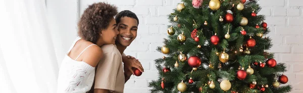 Mulher americana africana alegre com bola de Natal abraçando namorado perto de abeto decorado, bandeira — Fotografia de Stock