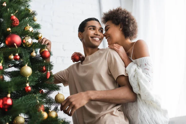 Young african american woman holding christmas ball near smiling boyfriend decorating pine tree — Stock Photo