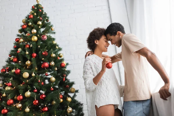 Mujer afroamericana feliz con bola de Navidad de pie cerca de novio feliz y abeto decorado - foto de stock