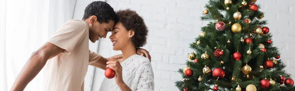 Vista lateral do alegre casal afro-americano com bola de Natal perto de pinheiros decorados, bandeira — Fotografia de Stock