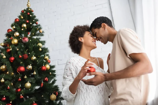Vista de ángulo bajo de la pareja afroamericana de pie cara a cara con los ojos cerrados cerca del árbol de Navidad en casa - foto de stock