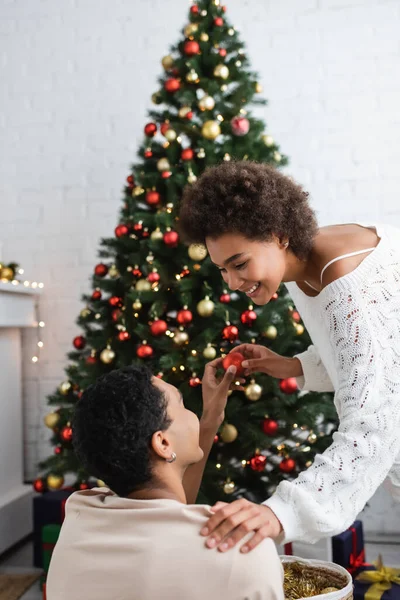 Heureuse femme afro-américaine en pull tricoté blanc prenant boule de Noël de petit ami près de pin — Photo de stock