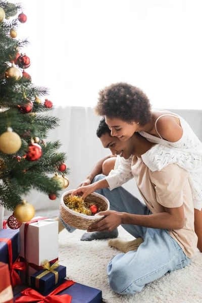 Heureux couple afro-américain tenant panier en osier avec des boules près de boîtes-cadeaux et arbre de Noël — Photo de stock