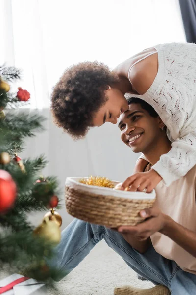 Joyeux homme afro-américain tenant panier en osier près de jeune copine décoration floue arbre de Noël — Photo de stock