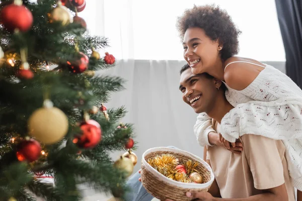 Alegre pareja afroamericana mirando decorado árbol de Navidad cerca de canasta de mimbre con bolas - foto de stock
