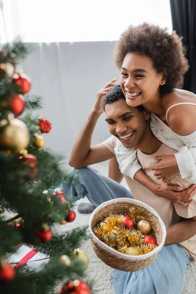 Mulher americana africana alegre abraçando namorado segurando bugigangas perto de árvore de Natal borrada — Fotografia de Stock