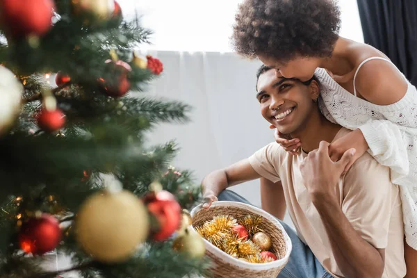 Curly african american woman hugging cheerful boyfriend holding wicker basket with baubles near christmas tree — Stock Photo