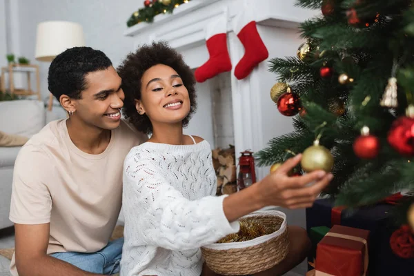 Sonriente mujer afroamericana en suéter calado blanco decorando pino cerca de novio feliz - foto de stock