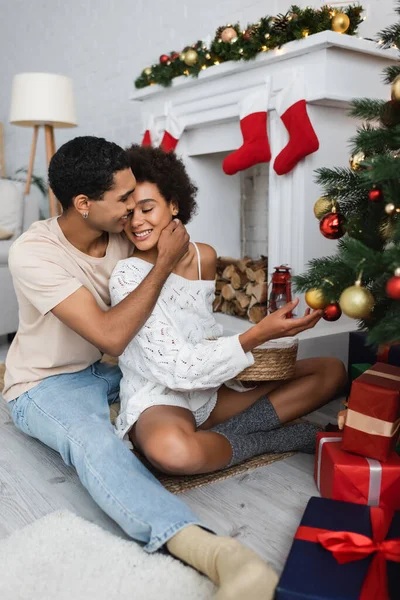 Feliz afroamericano hombre abrazando sexy novia sentado en el suelo cerca de la chimenea y el árbol de Navidad - foto de stock