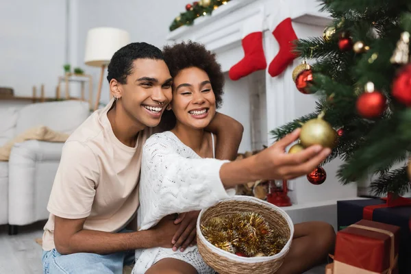 Mujer afroamericana feliz decoración árbol de Navidad cerca de novio y canasta de mimbre con oropel - foto de stock