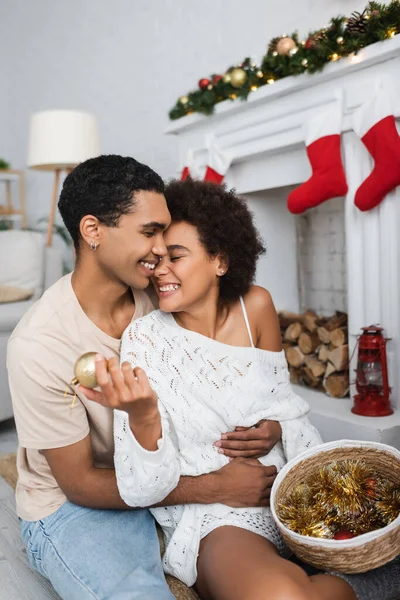 Heureux homme afro-américain embrassant petite amie tenant boule de Noël et tinsel dans le panier en osier — Photo de stock
