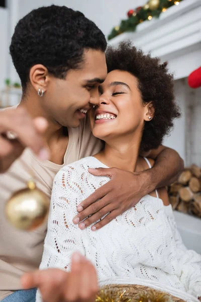 Alegre africano americano hombre abrazando sexy novia mientras celebración borrosa navidad bola - foto de stock