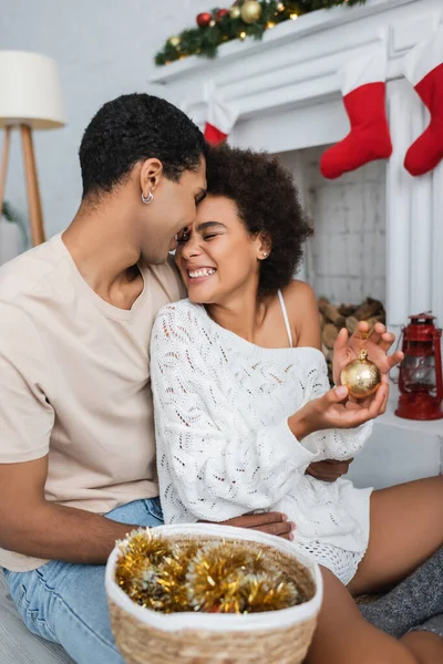 Aufgeregte Afroamerikanerin hält goldene Weihnachtskugel neben Freund mit Lametta im Weidenkorb — Stockfoto
