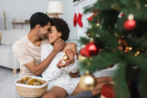Jeune homme afro-américain étreignant femme heureuse avec boule près flou arbre de Noël — Photo de stock