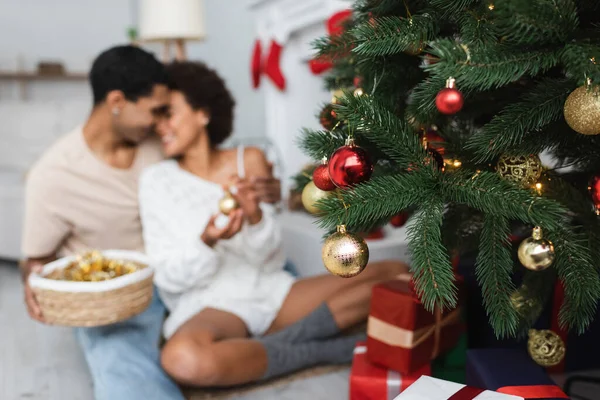 Enfoque selectivo de pino verde con bolas de Navidad brillantes cerca de pareja afroamericana sobre fondo borroso - foto de stock