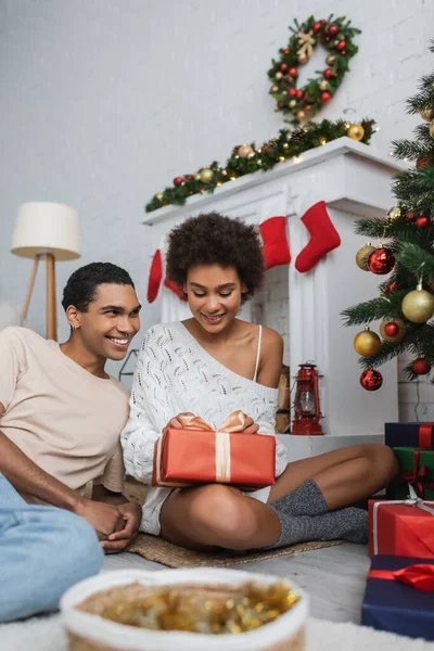 Happy and sexy african american woman sitting on floor with christmas present near fireplace and smiling boyfriend — Stock Photo