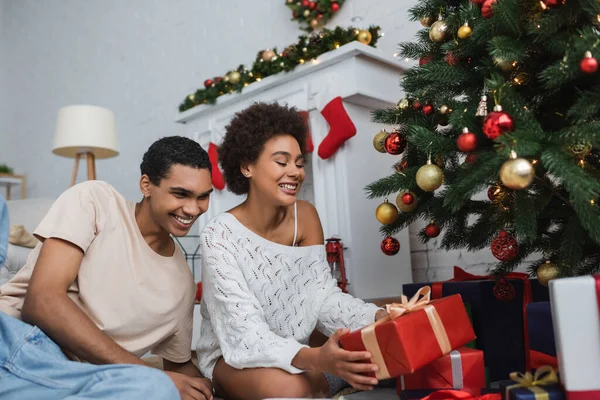 Mujer afroamericana feliz en suéter de punto blanco sosteniendo caja de regalo cerca de novio y árbol de Navidad - foto de stock