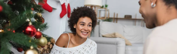 Cheerful african american woman looking at blurred boyfriend near christmas tree, banner — Stock Photo