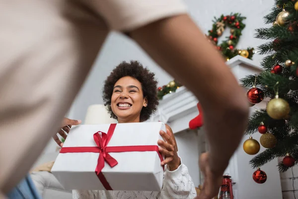 Low angle view of overjoyed african american woman holding large gift box near christmas tree and blurred boyfriend — Stock Photo