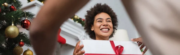 Low-Winkelaufnahme einer lachenden afrikanisch-amerikanischen Frau mit Weihnachtsgeschenk in der Nähe eines verschwommenen Freundes, Banner — Stockfoto