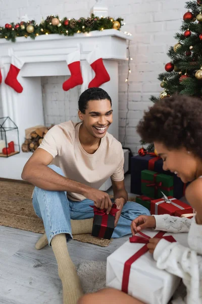 Sourire homme afro-américain regardant petite amie ouvrir boîte cadeau près de l'arbre de Noël — Photo de stock
