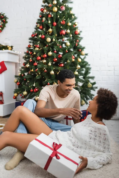 Joven afroamericano hombre sonriendo cerca de árbol de Navidad y sexy mujer sosteniendo caja de regalo en el suelo - foto de stock