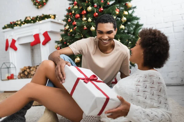 Alegre pareja afroamericana sosteniendo gran caja de regalo cerca borrosa árbol de Navidad en la sala de estar - foto de stock