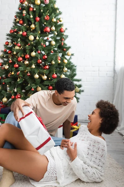 Young african american man holding gift box near christmas tree and laughing girlfriend — Stock Photo