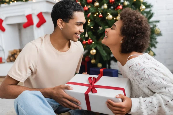Sonriente afroamericano hombre presentando regalo de navidad a novia complacida - foto de stock