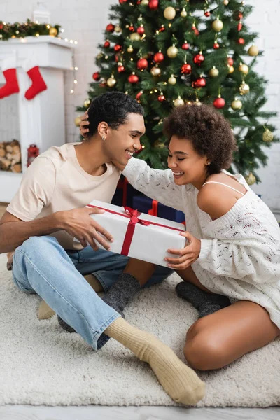 Alegre pareja afroamericana sentada en el suelo con caja de regalo cerca del árbol de Navidad - foto de stock