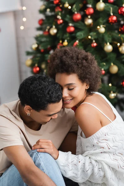 Sexy africana americana mujer sonriendo con los ojos cerrados cerca joven novio y borrosa árbol de Navidad - foto de stock