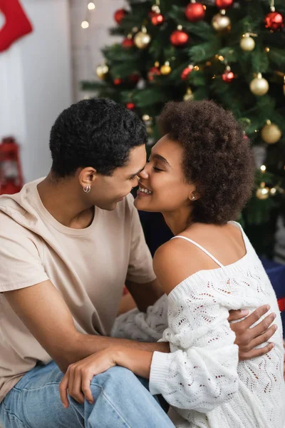 Young african american man embracing and kissing sexy woman in white knitted sweater near christmas tree — Stock Photo