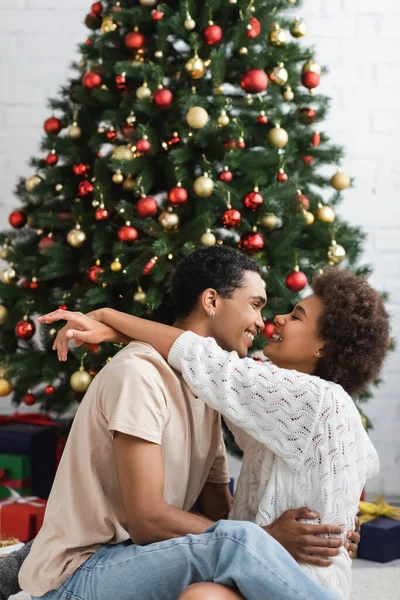 Seitenansicht eines jungen afrikanisch-amerikanischen Paares, das sich in der Nähe des Weihnachtsbaums umarmt und ansieht — Stockfoto